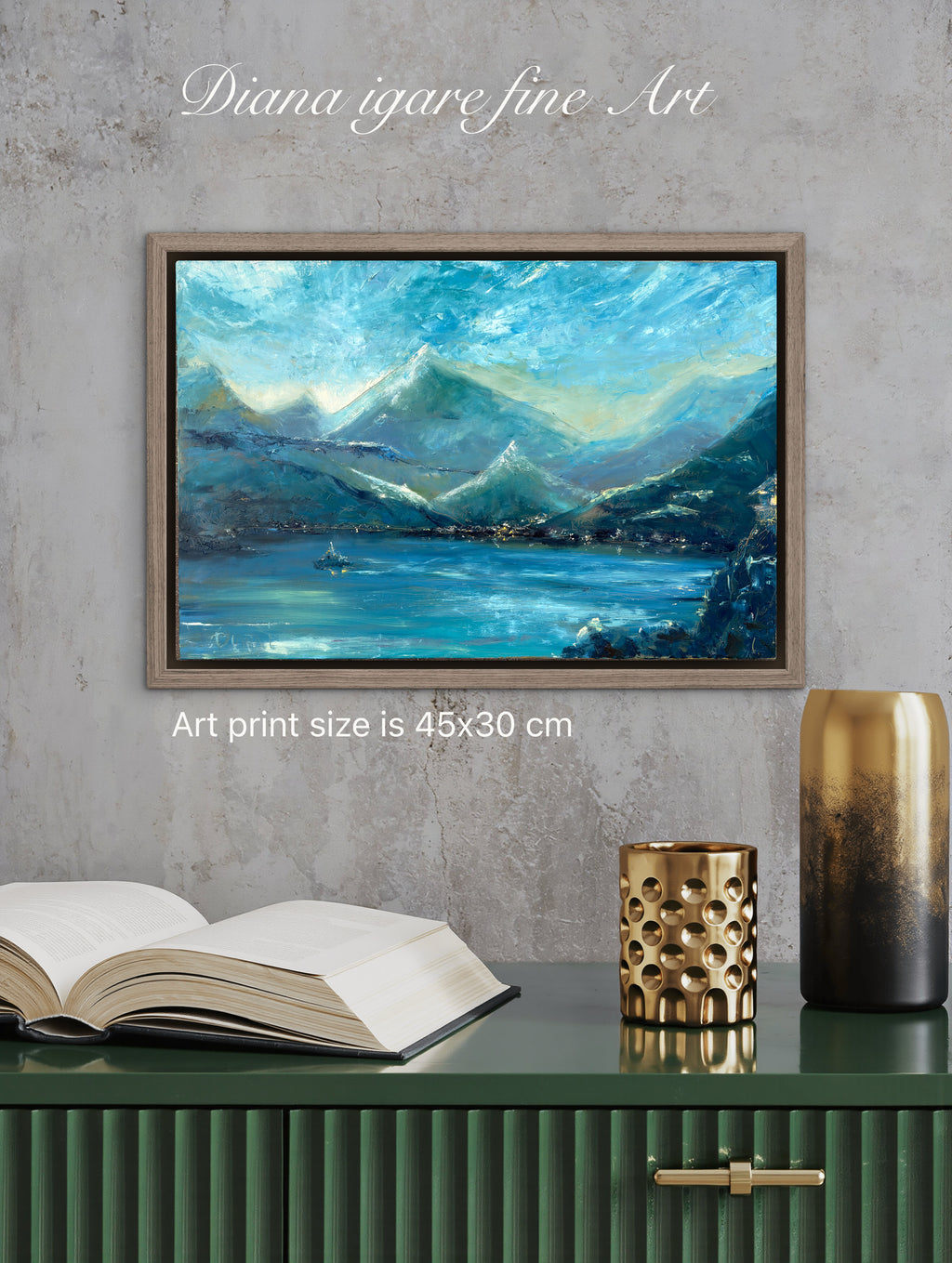 A framed landscape art print depicting blue mountains and a lake, displayed on a gray textured wall above a green cabinet with an open book and decorative vases. Text overlays read 'Diana igare fine Art' and 'Art print size is 45x30 cm'.
