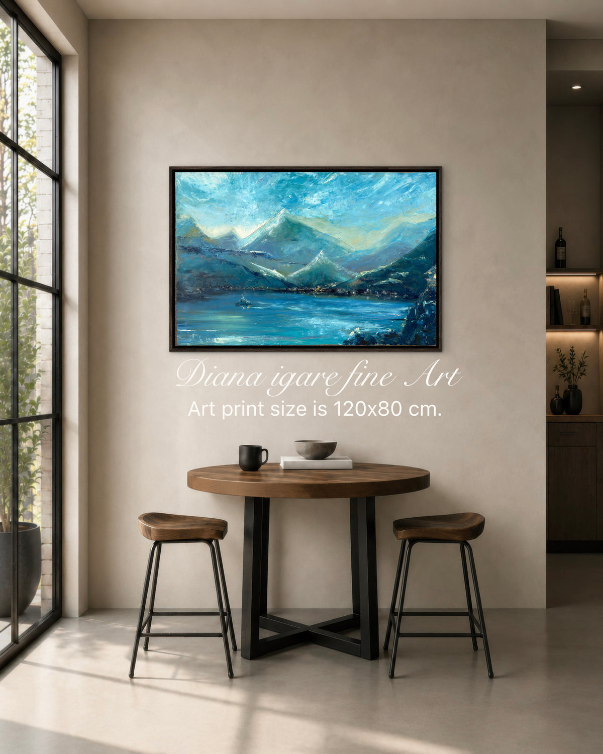 A framed landscape art print depicting blue mountains and a lake, displayed on a beige wall in a modern dining room setting with a wooden table and two stools. White text overlay reads 'Diana igare fine Art Art print size is 120x80 cm.