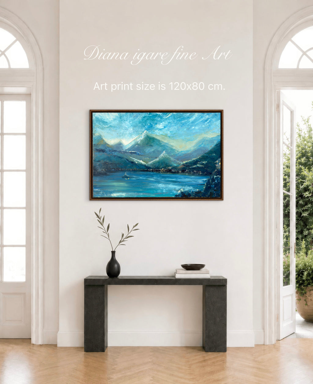 A framed landscape art print featuring blue mountains and a lake, displayed on a white wall above a dark console table in a bright room. Text on the wall reads 'Diana igare fine Art' and 'Art print size is 120x80 cm.